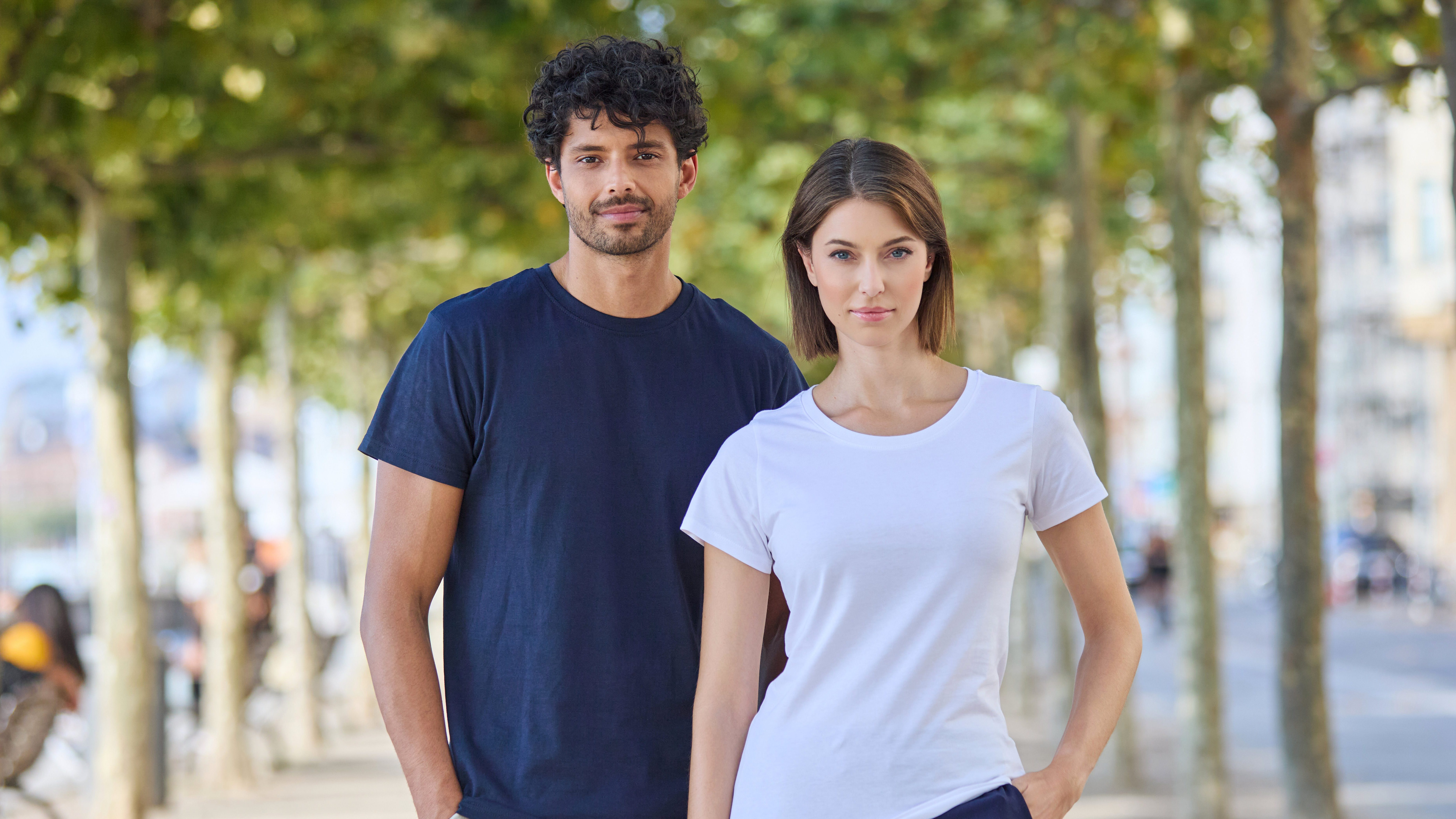 Ein Mann in dunkelblauem (Navy) T-Shirt und eine Frau in weißem T-Shirt stehen nebeneinander auf einer von Bäumen gesäumten Straße und tragen schlichte, zeitlose Basics.