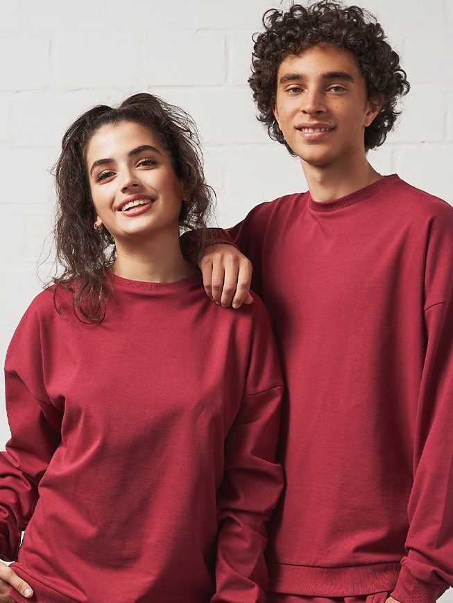 Zwei Personen (links Frau, rechts Mann, beide jung mit Locken) Mann lehnt mit einem arm auf der Schulter der Frau. Beide Tragen das "Oversized Sweatshirt" von wearecasual in der Farbe Berry. (dunkles leicht pinkes rot)